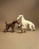 Three felt animal figurines, including a white dog, a brown dog, and a pug, arranged on a plain beige studio background.