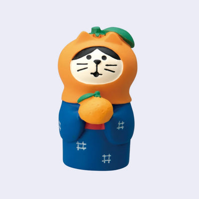 Small figure of a black and white cat wearing a blue kimono and holding a tangerine, while wearing a tangerine shaped hood hat.