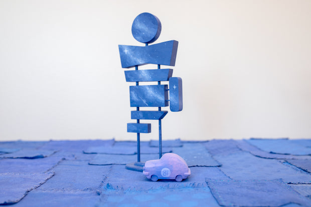 Pair of 2 carved wooden sculptures: a small retro looking purple car with 81 drawn on the side, and a large signpost with 7 different signs, though all painting like a dark blue night sky without any words.