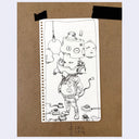 A hand-drawn black ink illustration on a white sheet of paper, taped to a brown cardboard background. The sketch shows a surreal character with a baby's head wearing a helmet and goggles, surrounded by small objects and baby faces.