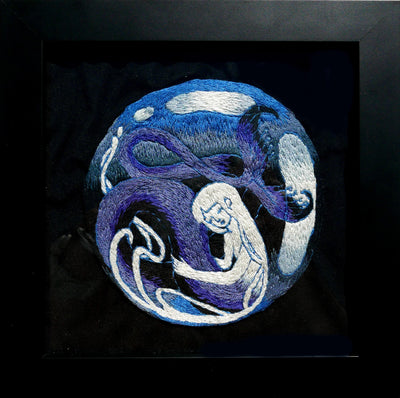 Blue, white and purple threaded hand embroidery of a mermaid with a long, winding tail. She hugs her tail.