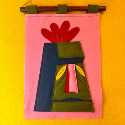 Colorful felt wall hanging with a design of a Tiki head with a red flower atop its head on a pink background against a yellow wall.