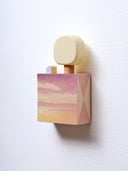 Small square wooden panel painted like an orange and purple sunset. Atop of it are small wooden sculpture: a tiny purple house and a rounded yellow tree.