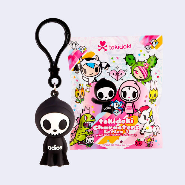 Small, soft PVC material bag charm of a skeleton, akin to the Grim Reaper with a simplistic face and cloaked body. Across its stomach reads "adios." It stands next to its product packaging.