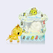 Small vinyl yellow unicorn figure with a fiery mane and a gold crown atop its head. It stands next to a clear plastic packaging that contains a squishy white unicorn inside.