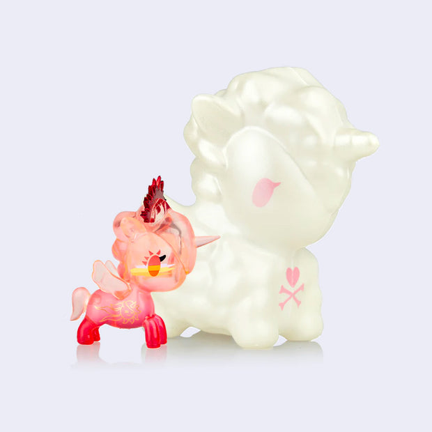 A small pink semi transparent unicorn figure with a red crown atop its head, standing next to a large squishy white unicorn.