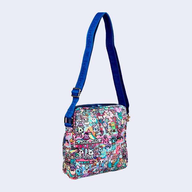 Small pouched shaped bag with an additional zipper section on the exterior. It has gold hardware details, and a cobalt blue sling strap attached. Bag is covered completely in a busy pastel color pattern featuring tokidoki characters with carnival food and drink imagery as well as carnival rides.