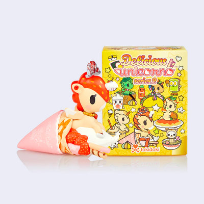 Vinyl unicorn with strawberry patterned mane, popping out of a strawberry crepe.  It stands next to its product packaging.