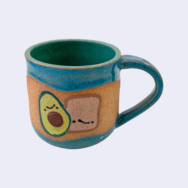 Ceramic mug with spotted finishing and an earthy brown exterior and bluish green interior. On the outside are painted on cartoon style avocado and toast, with simple expressions.