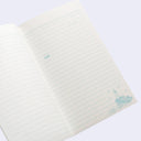 An open lined notebook with cream-colored pages and light blue printed text lines, featuring a small blue illustration of a lighthouse and birds in the bottom right corner.