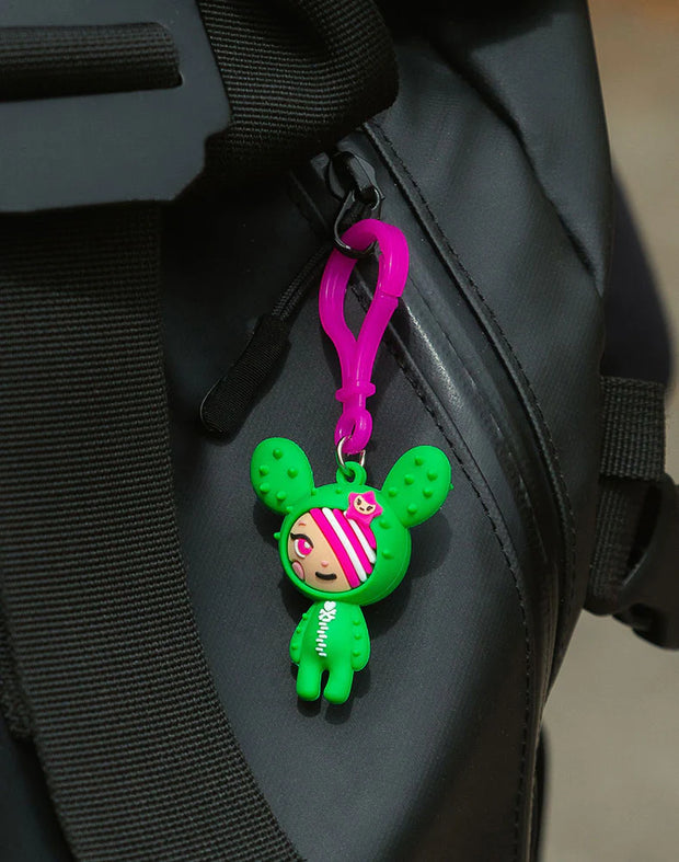 Small, soft PVC material bag charm of tokidoki's character SANDy, a small girl dressed in a green cactus costume. Keycharm is attached to a black backpack.