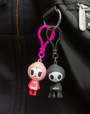 2 small, soft PVC material bag charms, attached to the same keyring on a black backpack. Charms are of tokidoki's characters: Ciao Ciao and Adios, 2 cute simplistic skeleton characters with their names written across their chests.