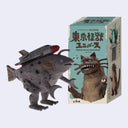 A toy monster figure with a fish-like body and a rocket on its back stands beside its illustrated product box. The box shows Japanese text and artwork for Tokyo Kaiju Universe.