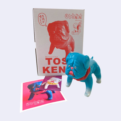 Teal blue soft vinyl figure of a pitbull type dog, with a red collar and white paws. It stands in front of its product packaging with a few notecard underneath its box.