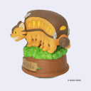 Wind-up music playing figurine of Catbus from My Neighbor Totoro, standing atop a greenery on a wooden looking base. Catbus is smiling with a slight curve in its stance.