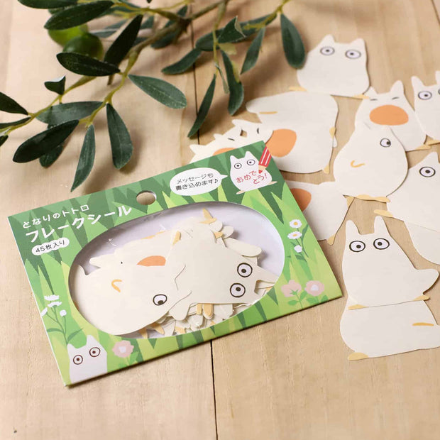 Packaged sticker sheet with cartoon chibi Totoro design on a wooden surface with green leaves.