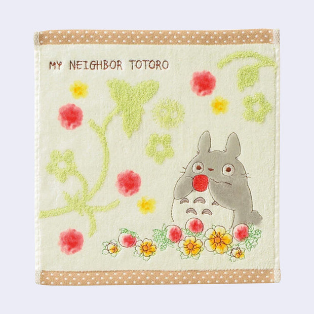 Square cream colored towel with brown with white polka dot border. A chibi Totoro sits amongst yellow and red flowers, and holds one up to its face. Floral patterns are in the background and "My Neighbor Totoro" is written across the top left.