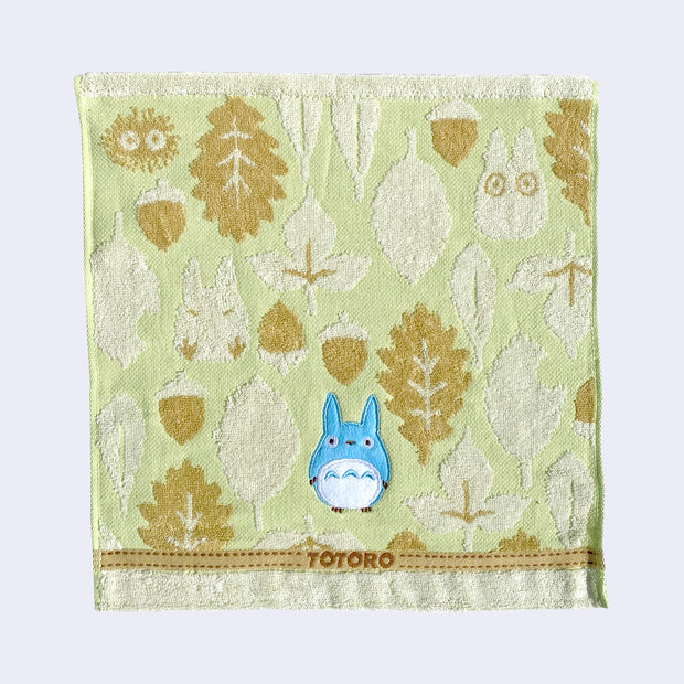 Light green square towel with a small blue embroidered Totoro in the bottom middle. Around are white and tan leaf and acorn patterns, with "Totoro" written small along the bottom.