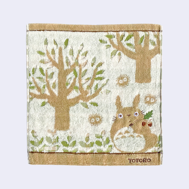 Light cream and tan square hand towel, with imagery of a small Totoro holding a branch with acorns. Dust sprites are around him and large trees with sparse leaves are in the background. 