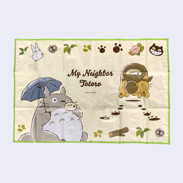 Polyproplyne mat, yellow background with a green outline featuring an illustration of Totoro playing a small wind instrument and holding an umbrella. Next to him, Catbus scurries away leaving paw prints. "My Neighbor Totoro" is written in the middle.