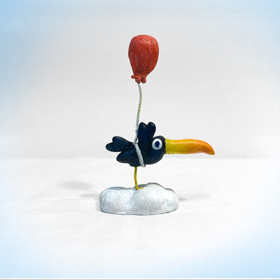 Sculpture of a cartoon style toucan, standing on a cloud. A single red balloon is tied around its body.