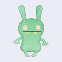 Plush doll, mostly flat in shape, of a bunny like character with wide set circular eyes and a straight lined mouth with 2 yellow round teeth peeking out.