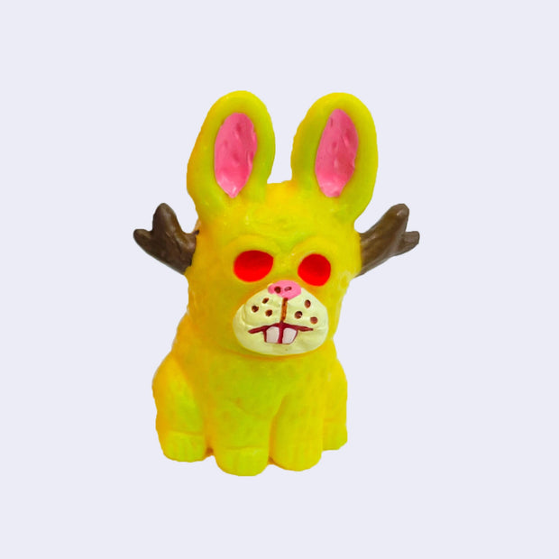 Vinyl figure of a yellow jackalope, sitting on the ground with hollowed red eyes and brown small antlers.
