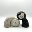 2 ceramic figures, leaning into one another. They have no arms or legs but cute faces and wear costumes with full hoods. One is a grayish white and the other is black.