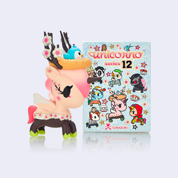 Vinyl unicorn figure, mainly pink with brown and green legs. Around its waist is a row of cherry blossoms and atop its head is a nest with a small bluebird in it and some branches. It stands next to its product packaging.