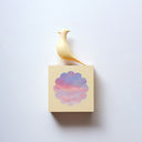 Small square wood panel with a purple and pink sunset shown within a flower outline, the rest of the panel is painted yellow. Atop of it is a carved wooden sculpture of a yellow bird.