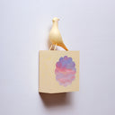 Small square wood panel with a purple and pink sunset shown within a flower outline, the rest of the panel is painted yellow. Atop of it is a carved wooden sculpture of a yellow bird.