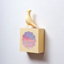Small square wood panel with a purple and pink sunset shown within a flower outline, the rest of the panel is painted yellow. Atop of it is a carved wooden sculpture of a yellow bird.