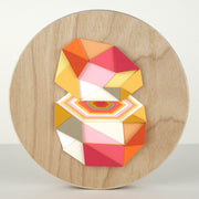 Geometric designed layered cut paper sculpture mounted on wood, creating a three dimensionality. A 10 sided shape is cut horizontally, revealing a hexagonal interior. Colors are orange, grey, yellow, pink and white.