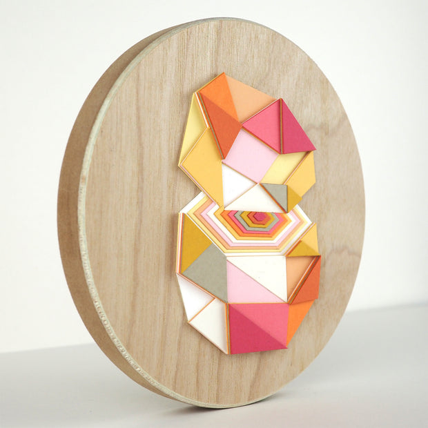 Geometric designed layered cut paper sculpture mounted on wood, creating a three dimensionality. A 10 sided shape is cut horizontally, revealing a hexagonal interior. Colors are orange, grey, yellow, pink and white.
