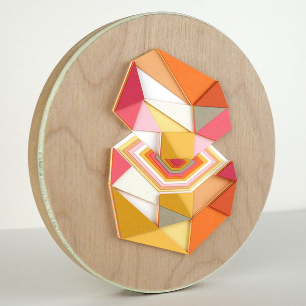 Geometric designed layered cut paper sculpture mounted on wood, creating a three dimensionality. A 10 sided shape is cut horizontally, revealing a hexagonal interior. Colors are orange, grey, yellow, pink and white.