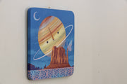 Painting of Saturn, with a simple cartoon face. It hangs largely behind a rocky bluff with a large butterfly resting atop them. A small row of breeze blocks lines the bottom of the piece.