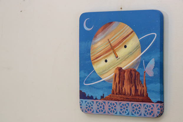 Painting of Saturn, with a simple cartoon face. It hangs largely behind a rocky bluff with a large butterfly resting atop them. A small row of breeze blocks lines the bottom of the piece.