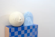 Small square wood panel painted with a blue and gray checkered patter. Atop of it are carved wooden sculptures: 2 blue mountainous rocks and a white sphere with a closed eye, calm smiling expression.