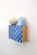 Small square wood panel painted with a blue and gray checkered patter. Atop of it are carved wooden sculptures: 2 blue mountainous rocks and a white sphere with a closed eye, calm smiling expression.