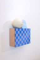 Small square wood panel painted with a blue and gray checkered patter. Atop of it are carved wooden sculptures: 2 blue mountainous rocks and a white sphere with a closed eye, calm smiling expression.