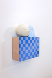 Small square wood panel painted with a blue and gray checkered patter. Atop of it are carved wooden sculptures: 2 blue mountainous rocks and a white sphere with a closed eye, calm smiling expression.