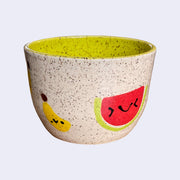 Ceramic bowl with spotted finishing and an earthy cream exterior and lime green interior. On the outside are painted on cartoon style banana and watermelon, with simple expressions.
