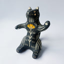 Black ceramic sculpture with cream colored carved design of various lines around its body and an orange poppy flower on its stomach. It sits on its behind and has one arm up as though waving.