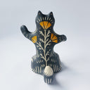 Black ceramic sculpture with cream colored carved design of various lines around its body and an orange poppy flower on its stomach. It sits on its behind and has one arm up as though waving.