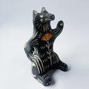 Black ceramic sculpture with cream colored carved design of various lines around its body and an orange poppy flower on its stomach. It sits on its behind and has one arm up as though waving.