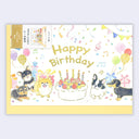 A square birthday card with a pale yellow envelope , decorated with colorful illustrations of small dogs, a birthday cake with candles, balloons, confetti, musical notes, and the words 'Happy Birthday.' A small package insert is visible along the left edge.