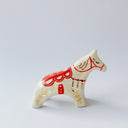 Cream colored ceramic sculpture of a white doll-like horse, with gold patterning on it its body and for its mane and a red ornate saddle.