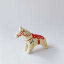 Cream colored ceramic sculpture of a white doll-like horse, with gold patterning on it its body and for its mane and a red ornate saddle.