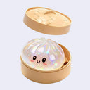 A cute bun-shaped squishy toy with a smiling face, set inside a round beige container with a matching lid shown floating above. The squishy is made of iridescent glittery plastic.