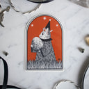 Sticker of a cat holding a large egg and standing in grass, dressed as a wizard. Background is orange.